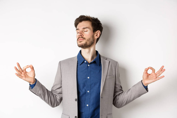 Calm and focused businessman meditating with eyes closed and hands spread sideways, finding peace in meditation, practice yoga breathing, standing on white background - Photo, Image