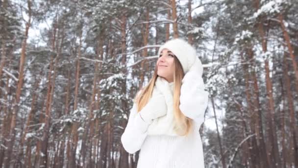 Όμορφο χαμογελαστό κορίτσι στο χειμερινό δάσος. Ποζάροντας σε ένα λευκό πουλόβερ, καπέλο και γάντια - Πλάνα, βίντεο