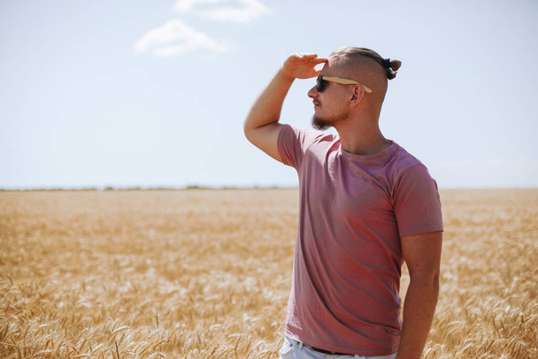 barbudo chico en rosa camiseta y gafas de sol mira a la distancia en un campo de trigo amarillo dorado - Foto, Imagen