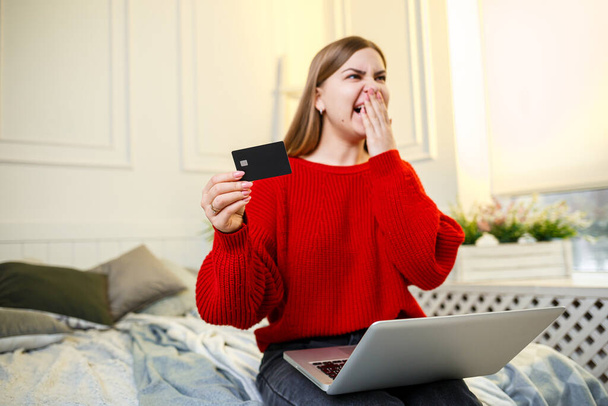 Una mujer sostiene una tarjeta de crédito en sus manos, lista para comprar, vistiendo un suéter rojo, sentada en la cama en casa, un cliente del banco mostrando su tarjeta de crédito. Compras en línea desde casa. Enfoque selectivo - Foto, Imagen