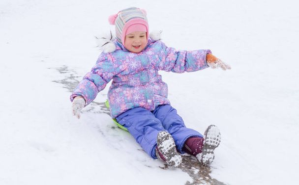 A little pretty girl in a purple overalls slides down an ice slide, winter children's games, a child in the park in winter. - Photo, Image