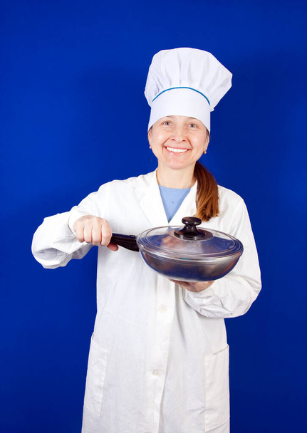 Senior-Köchin in Haube mit Edelstahl-Pfanne vor blauem Hintergrund - Foto, Bild