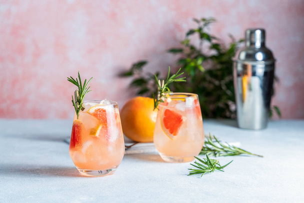 Tequila-Cocktail oder kalte Limonade mit Grapefruitsaft, getönt mit dem Aroma eines frischen Rosmarinzweiges auf pastellrosa Hintergrund. - Foto, Bild