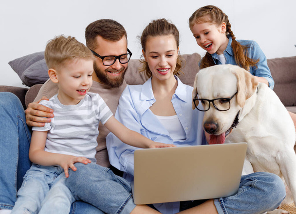 Fröhliche Familie mit kleinen Kindern und lustigem Labrador Retriever Hund in Brille, der sich um Laptop versammelt und interessante Filme anschaut, während er die Freizeit zu Hause zusammen verbringt - Foto, Bild