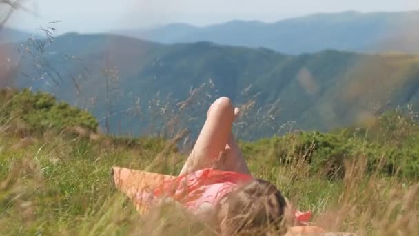 Junge Frau mit langen Beinen legt sich auf grüne Wiese an einem warmen, sonnigen Tag in den sommerlichen Bergen und genießt die Aussicht auf die Natur. - Filmmaterial, Video