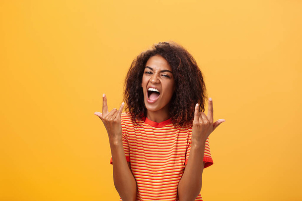 Знімок приголомшеного радісного стильного африканського кольору жінка з Америки почуває благоговійний потрясіння на вечірці крики від радості і задоволення, показуючи рок-н-рол жест, що ставить над помаранчевим фоном - Фото, зображення