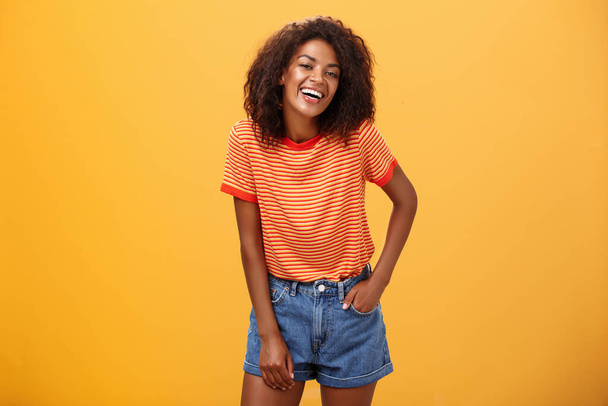 Retrato de la carismática encantadora afroamericana elegante afroamericana mujer en pantalones cortos de moda y camiseta riendo felizmente disfrutando hablando con gente fresca riendo posando sobre fondo naranja - Foto, Imagen