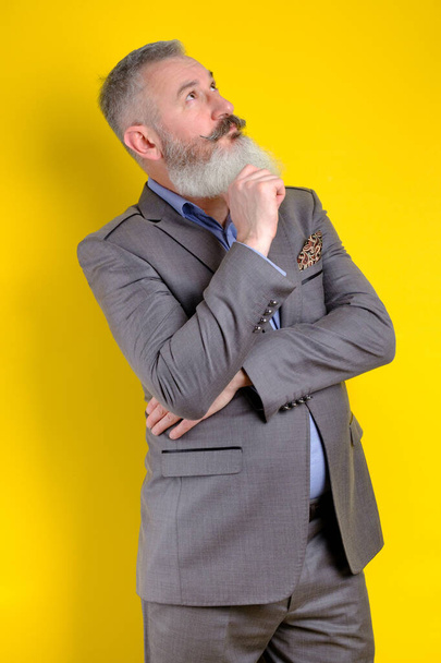 Studio portrait mature bearded man dressed in gray business suit looks pensively aside, thinks about something, yellow background - Photo, Image