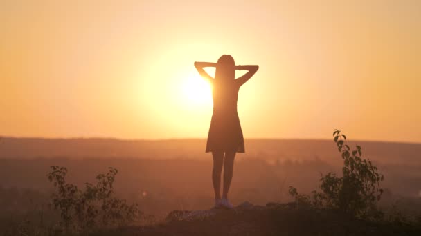 Молода жінка в літній сукні, що стоїть на відкритому повітрі з витягнутими руками, насолоджується видом на яскравий жовтий захід сонця
. - Кадри, відео