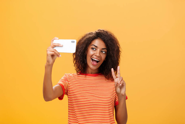 Χαμογέλα στην κάμερα. Ελκυστική και κομψή αυτοπεποίθηση σκούρο δέρμα θηλυκό μοντέλο με σγουρά μαλλιά δείχνει χειρονομία ειρήνης, ενώ λαμβάνοντας selfie κρατώντας smartphone κοντά στο πρόσωπο και χαμογελώντας στην οθόνη gadget - Φωτογραφία, εικόνα