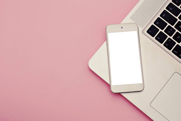 Mockup-Bild eines weißen Mobiltelefons mit leerem weißen Bildschirm mit modernem rosa Bürotisch mit Laptop zur Eingabe des Textes im Kopierraum Draufsicht, flache Lage. - Foto, Bild