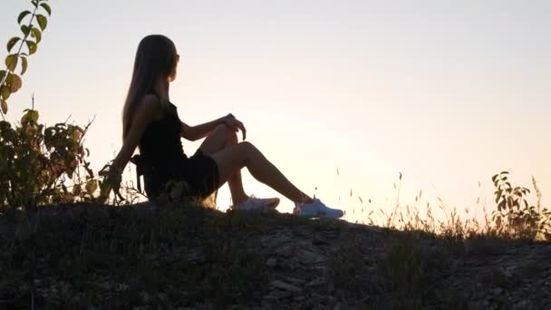 Jeune femme élégante en robe courte noire assise sur un rocher relaxant en plein air le soir d'été. Femme à la mode profitant d'un coucher de soleil chaud dans la nature. - Séquence, vidéo