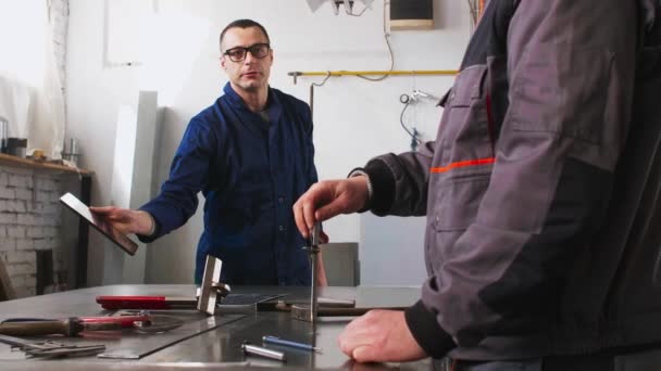 Джинсовий козел вимірює шматок олова у своїй майстерні, і він бере інструкцію від колеги-інженера, який тримає планшет
. - Кадри, відео