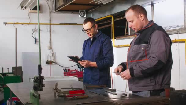 Een tinsmid en zijn collega-ingenieur werken samen aan een werktaak in hun werkplaats, terwijl de ingenieur tablet gebruikt.. - Video