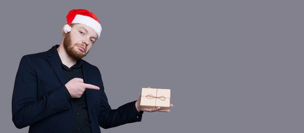 Uomo barbuto con cappello di Babbo Natale indossa abbigliamento formale sta indicando il presente in mano in posa su un muro grigio vicino allo spazio libero - Foto, immagini