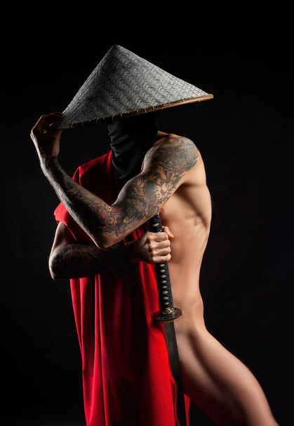 ein nackter schlanker Mann Samurai in rotem Umhang und asiatischem Hut mit tätowierter Katana - Foto, Bild
