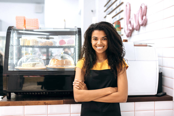 Amable chica afroamericana, camarera o propietaria de un negocio, se encuentra en un café cerca de los postres, con los brazos cruzados, se ve y sonríe a la cámara - Foto, Imagen