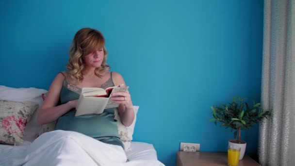 Een zwangere vrouw in een semi-zittende positie in haar bed en ze ziet er gelukkig uit terwijl ze een boek leest. Ze heeft één hand op haar buik.. - Video