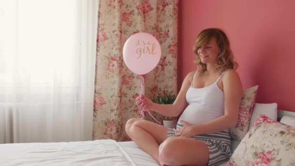Een gelukkige zwangere blanke vrouw in haar bed met een ballon in haar hand terwijl de inscriptie "het is een meisje. Ze houdt één hand op haar buik.. - Video