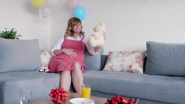 Egy mosolygó és boldog terhes fehér nő ül a nappaliban, miközben apró babaruhákat néz.. - Felvétel, videó