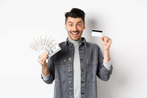 Opgewonden man met plastic credit card en dollarbiljetten, staande geamuseerd op witte achtergrond - Foto, afbeelding