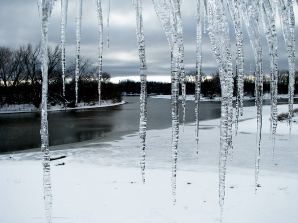 Зимний вид на реку и сосульки через окно. Сосульки, свисающие с крыши, зимний день. - Фото, изображение