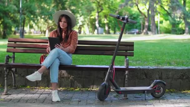 帽子を被った若い女性が公園のベンチに座って、タブレットを手にスクロールしている。電動スクーターは彼女の隣に駐車されていますが、木と緑の色が背景に優勢です。. - 映像、動画