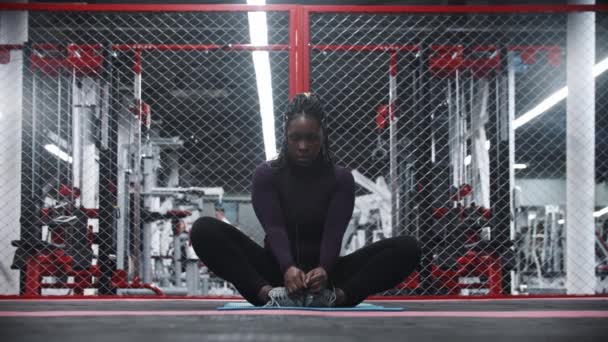 Μια Αφροαμερικανή που κάθεται στο στρώμα γιόγκα στο γυμναστήριο και γέρνει προς τα εμπρός. - Πλάνα, βίντεο