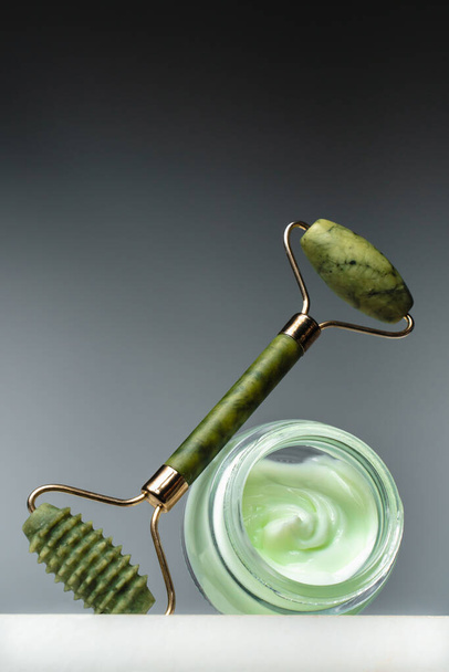 Crema facial y rodillo de masaje de jade en un pedestal. Elevación para una piel sana y juvenil. Cosmética ecológica sobre fondo negro. Cuidado de la piel tendencia - Foto, imagen