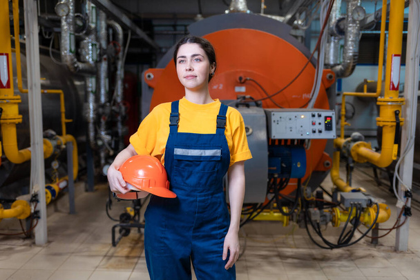 Das Konzept der industriellen Fertigung. Eine junge lächelnde Frau in Uniform hält einen Schutzhelm in der Hand und posiert im Heizungsraum. - Foto, Bild