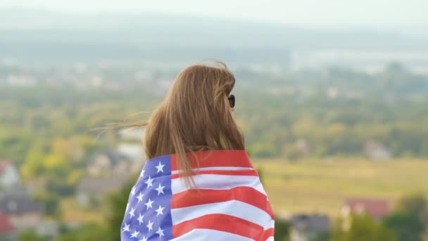 Молодая счастливая женщина с длинными волосами, держа махать на ветру американский национальный флаг на ее sholders отдыхает на открытом воздухе наслаждаясь теплым летним днем. - Кадры, видео
