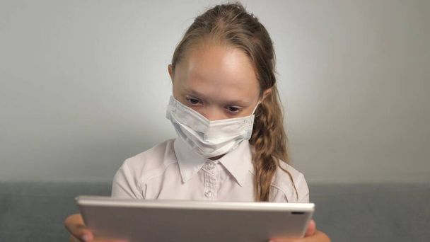 A child in mask plays on his tablet in the room on couch. A girl in a protective mask during an online lesson. A student in medical mask teaches homework at home with tablet. Quarantine, viral danger - Photo, Image