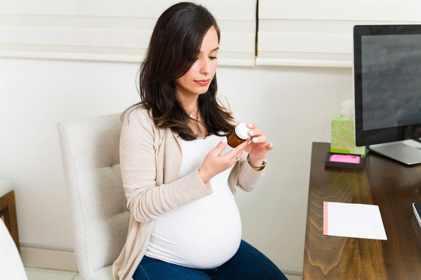 Hermosa madre embarazada leyendo la etiqueta y las instrucciones de un frasco de píldoras recetadas mientras está sentada en el consultorio del médico  - Foto, Imagen