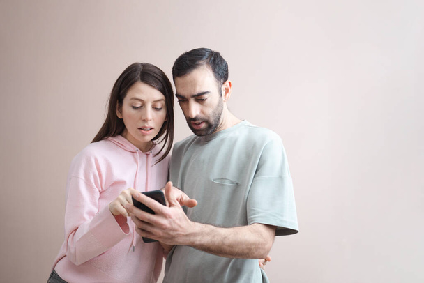 Zwei erwachsene Männer und eine erwachsene Frau schauen schockiert auf den Handybildschirm. Online einkaufen, Spaß haben. In lässigem pastellrosa Kapuzenpulli. Leerer Hintergrund, Kopierplatz für Text, Studioaufnahme, Nahaufnahme - Foto, Bild