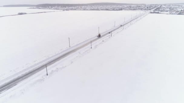 Luftaufnahme eines gelben Traktors mit Schaufeln auf der schneebedeckten Landstraße durch die Felder. Strommasten und Drähte am Straßenrand. Autos fahren die Straße entlang. Bewölkter Wintertag - Filmmaterial, Video