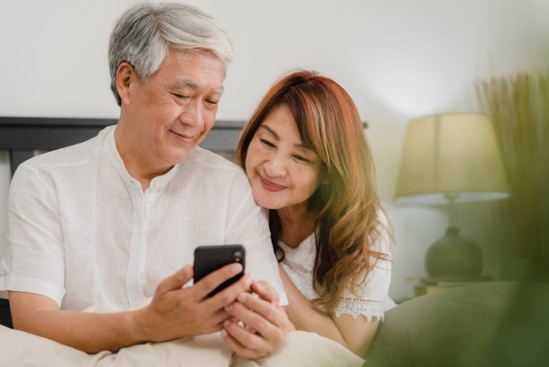 Asian senior couple using mobile phone at home. Asian Senior Chinese grandparents, husband and wife happy after wake up, watching movie lying on bed in bedroom at home in the morning concept. - Fotografie, Obrázek