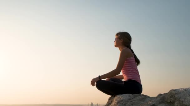 Entspannte junge Frau sitzt draußen auf einem großen Stein und genießt den warmen Sommertag. Mädchen meditieren und entspannen bei Sonnenuntergang in der Natur. - Filmmaterial, Video