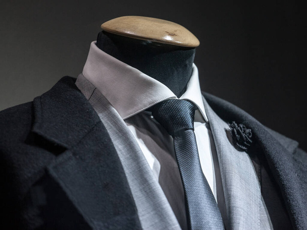 Men formal Suit, with a white shirt, a black tie, and a black lapel flower pin and grey jacket on display on dummy in front of a tailor store, on a window. It is one of the main formal garments in male fashion - Photo, Image