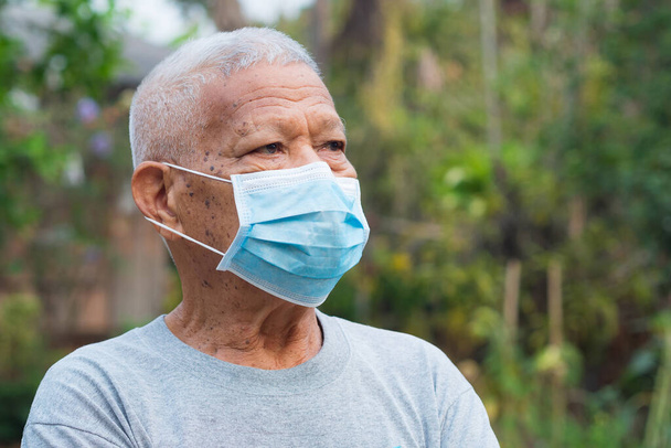Ένα πορτραίτο ενός ηλικιωμένου άνδρα με μάσκα προσώπου που κοιτάζει αλλού ενώ στέκεται σε έναν κήπο. Μάσκα για την προστασία του ιού, covid-19, coronavirus, βακτήρια, και πολλά άλλα. Έννοια των ηλικιωμένων και της υγειονομικής περίθαλψης. - Φωτογραφία, εικόνα