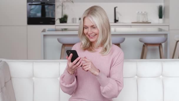 Excited happy mature old 60s woman winner holding smartphone using mobile app winning online lottery bid, celebrating success, making Yes gesture, while sitting on couch at home. - Footage, Video
