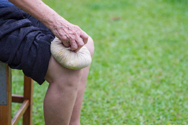 Een oudere vrouw gebruikt een Thaise kruidenkompres bal op de knie. Kniepijn kan veroorzaakt worden door spierspanning, tendinose, artrose (OA). Ruimte voor tekst. Begrip ouderen en gezondheidszorg. - Foto, afbeelding