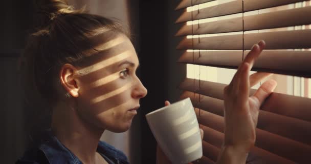 vrouw kijkt door het raam blinds in het zonlicht en het drinken van koffie - Video