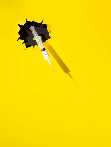 Jeringa irrumpiendo a través de un agujero negro sobre fondo amarillo. Concepto de vacuna Coronavirus mínimo o 2019-nCoV o COVID-19.  - Foto, imagen