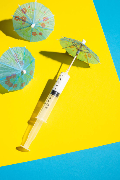 Jeringa con paraguas de cóctel en la playa hecha de papel amarillo y azul. Verano 2021 turismo y concepto de viaje. Escena mínima de Coronavirus o 2019-nCoV o COVID-19.  - Foto, imagen