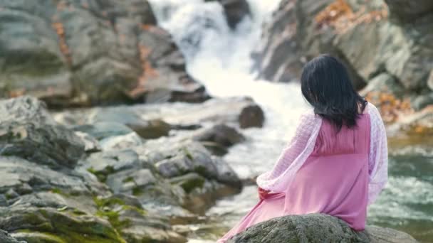 Una mujer de pelo negro en vestido de noche largo de moda sentado en una piedra cerca de un pequeño río de montaña con agua en movimiento rápido. - Metraje, vídeo