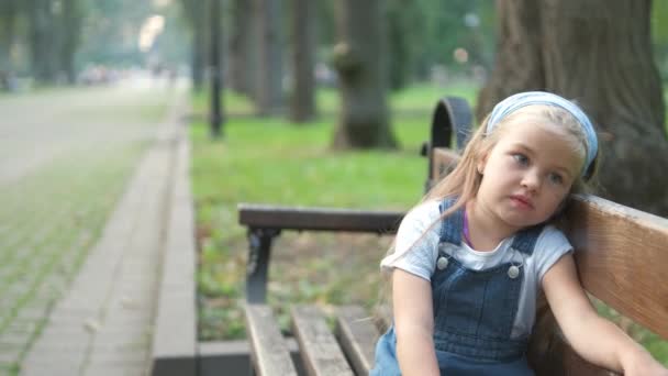 Mała poważna dziewczynka siedzi sama na ławce w letnim parku. - Materiał filmowy, wideo