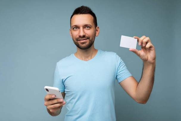 Fešák muž na sobě každodenní oblečení izolované na pozadí zdi držení a používání telefonu a kreditní karty platby při pohledu do kamery - Fotografie, Obrázek