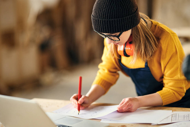 Εστιασμένη νεαρή ειδικευμένη τεχνίτρια που κρατάει σημειώσεις σε χαρτιά ενώ στέκεται στο τραπέζι με όργανα laptop και ξυλουργικής στο εργαστήριο - Φωτογραφία, εικόνα