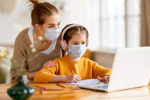 Jonge vrouw helpt dochtertje met medisch masker en koptelefoon tijdens les op afstand door coronavirus pandemie thuis - Foto, afbeelding