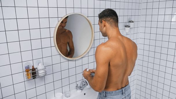 молодой мужчина без рубашки держит дезодорант возле зеркала в ванной комнате - Фото, изображение